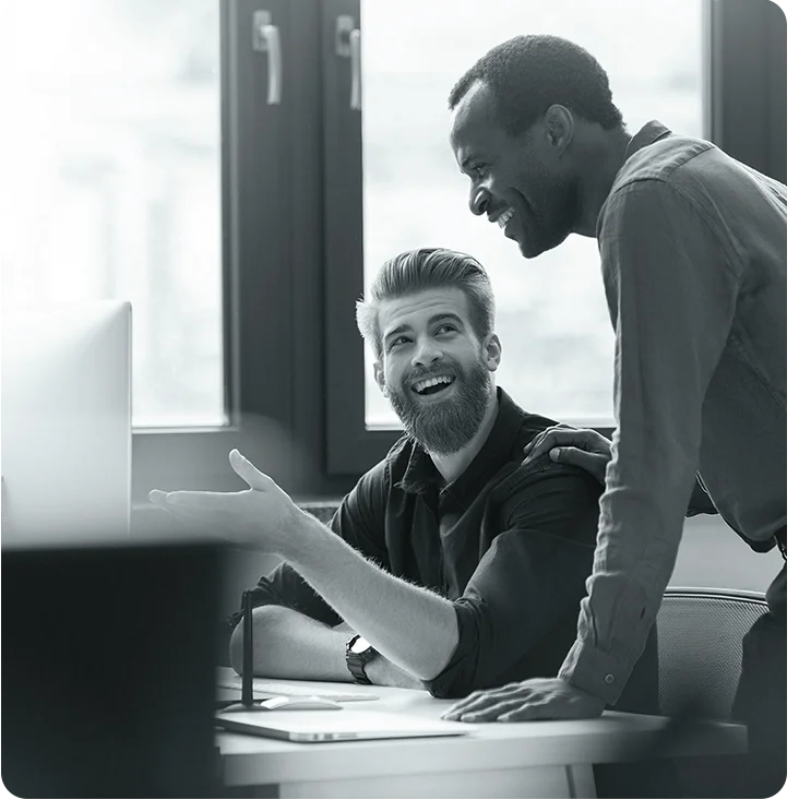 Imagem de dois homens em um escritório, sorrindo. Um homem está sentado mostrando uma tela para um homem sorridente com as mãos apoiadas sobre a mesa.